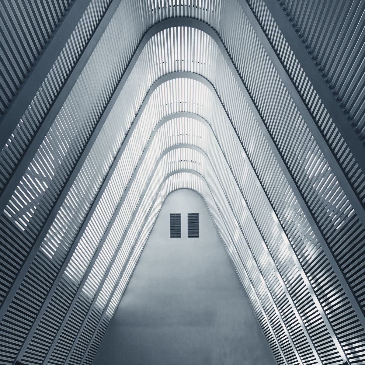 Looking up a tall atrium: pale ribbed balconies narrow to a skylight, in a cool blue-grey light.