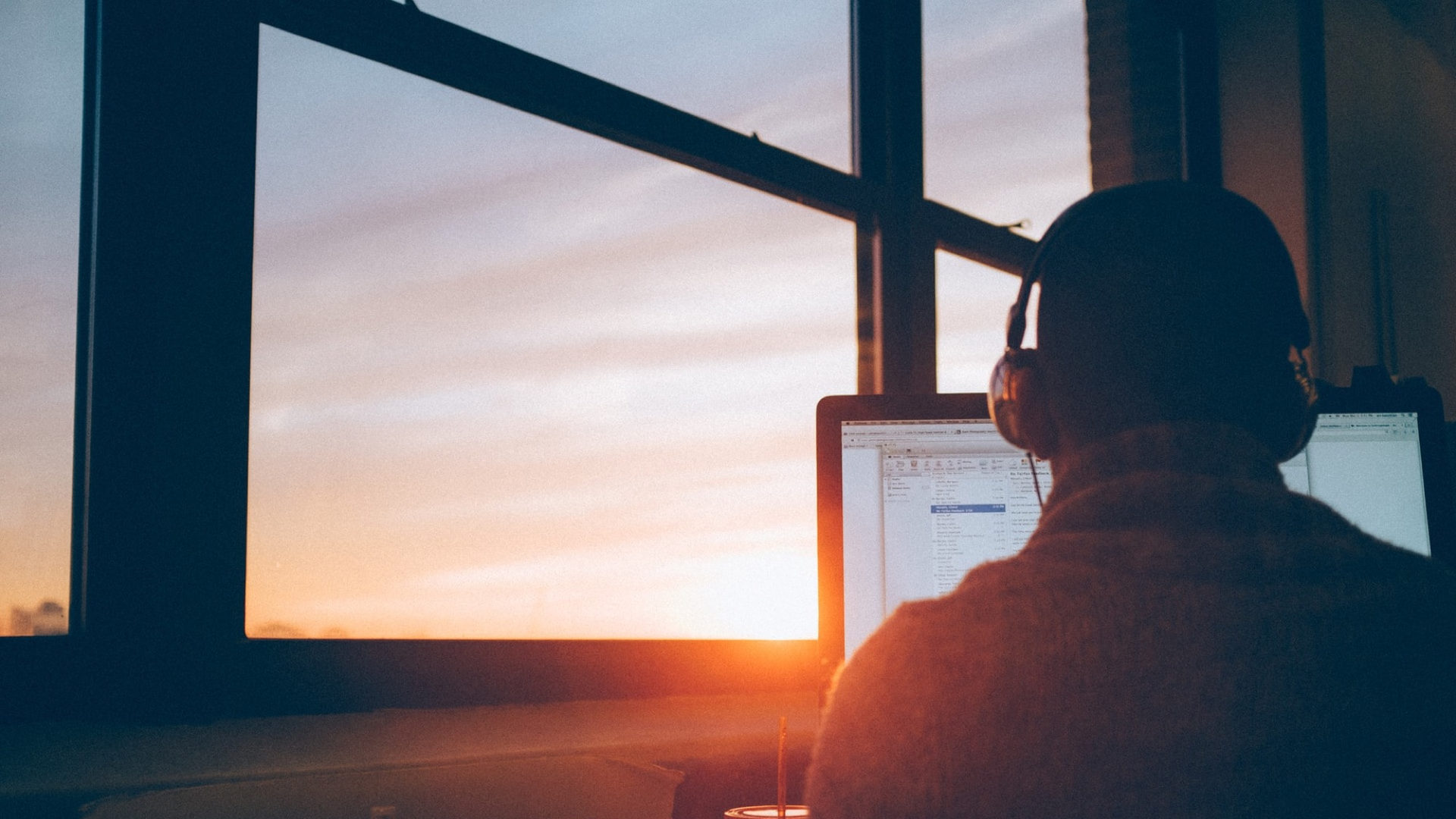 A person in headphones working at a monitor as the sun sets outside the window