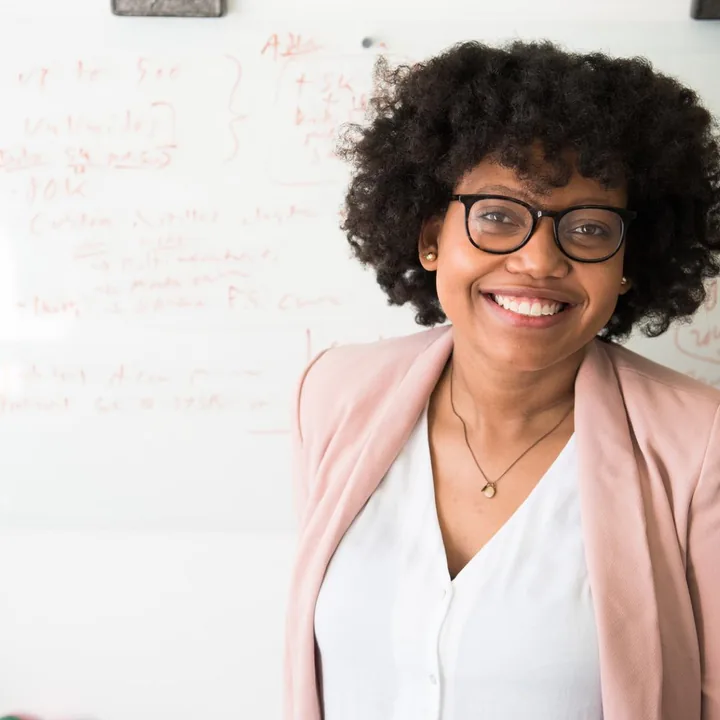 A smiling woman in glasses and a pink blazer, standing in front of a whiteboard