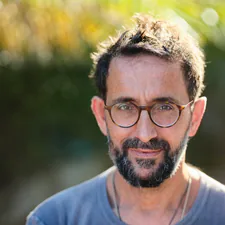 A bearded man in round glasses and a grey T-shirt, photographed outdoors
