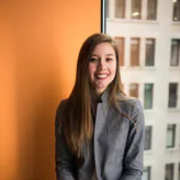 A smiling woman in a grey blazer, standing by an office window